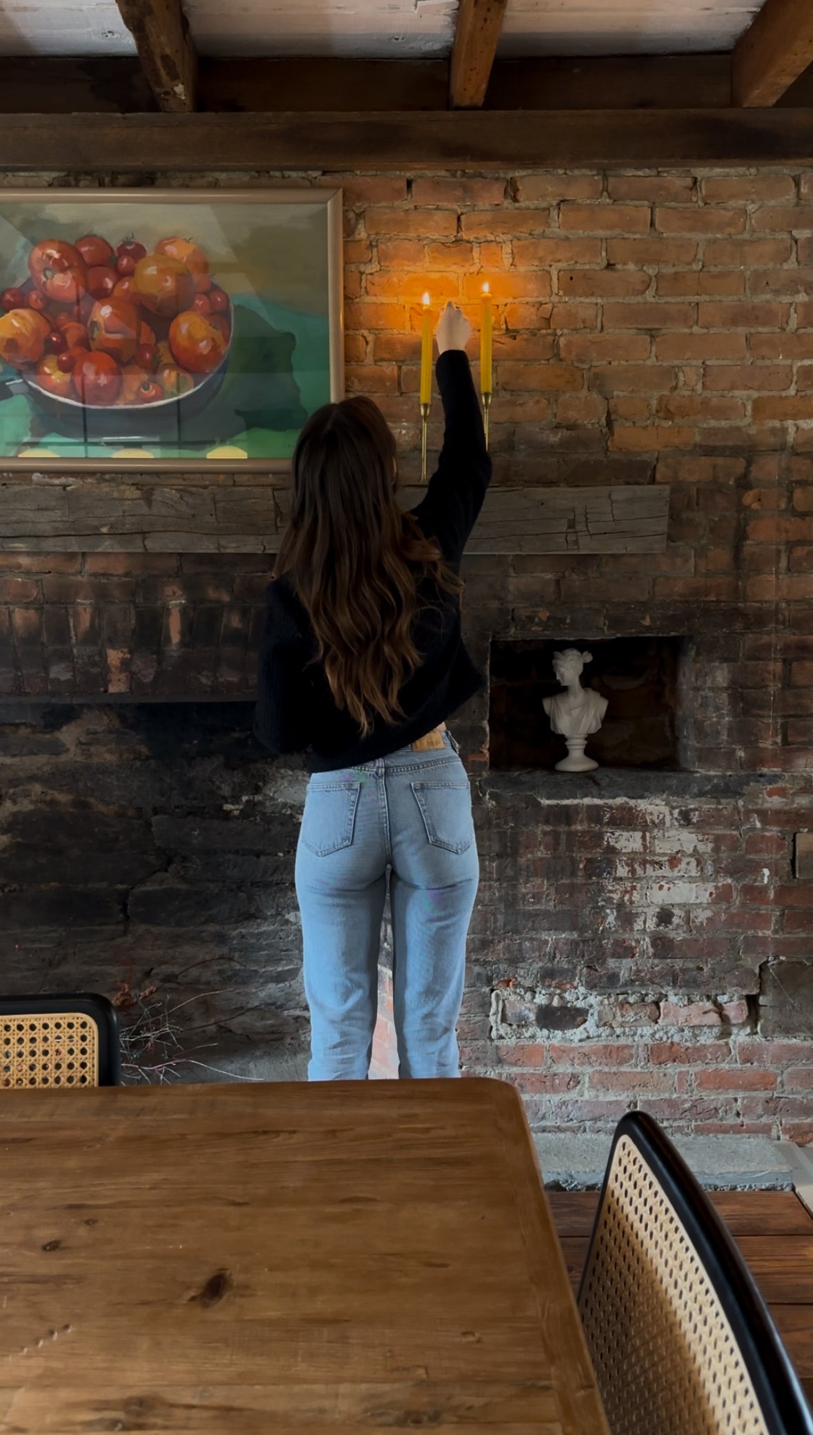 Person adjusting a light fixture on a brick wall with a painting of apples in the background.